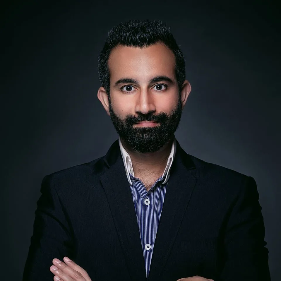 Image of Reemal standing with arms crossed in a black suit and. blue shirt. He has a beard.