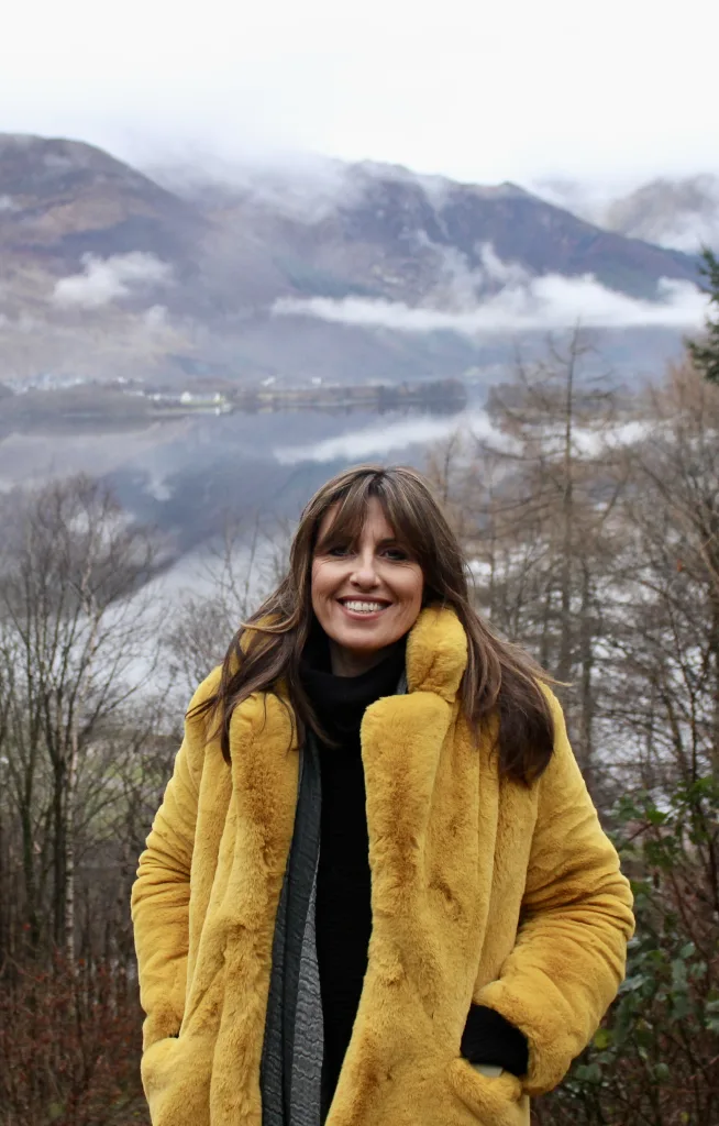 A woman in a yellow fur coat stands in front of a lake among cloud-covered mountains.
